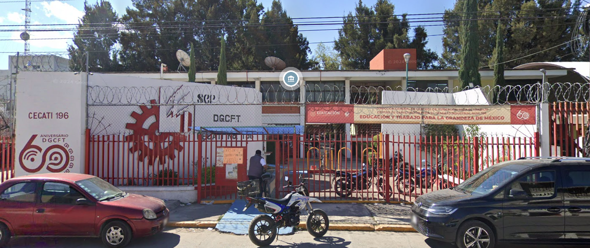 Fotografía en la que se muestra la entrada del plantel, conformada por una reja de color rojo que da acceso a las instalaciones. Del lado izquierdo de la rej<a hay un muro blanco, que en letras rojas dice CECATI 196.